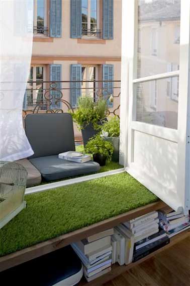 Un balcon étroit aménagé au centimètre près avec un revêtement de sol en gazon synthétique. Des coussins de sol alignées contre le sol et la rambarde exploitent l'espace extérieur au maximum