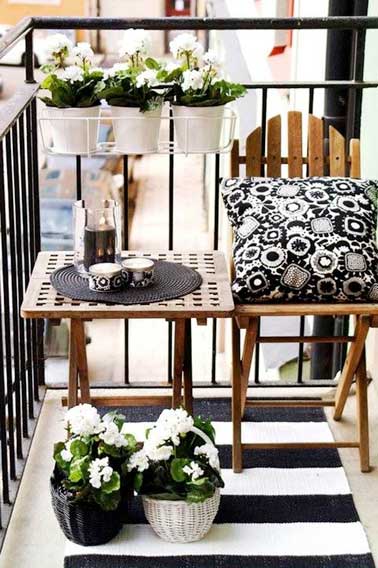 Un angle de balcon au carré avec table de jardin et chaise en bois alignés contre la balustrade. En symétrie, jardinières suspendus, paniers en osiers et tapis de sol à rayures noirs et blancs 