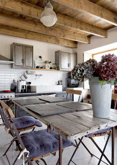Une cuisine de maison de vacances rénovée avec du carrelage métro. Table en bois, plafond poutre et meubles patinés amènent une note industrielle dans la pièce. 