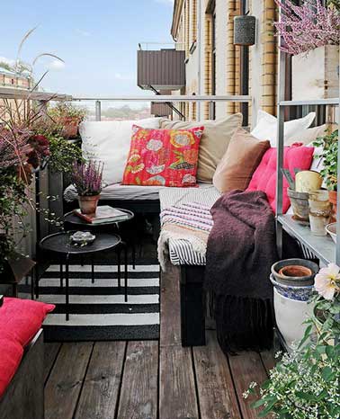 Un angle de balcon aménagé en coin cosy à l’aide d’une banquette recouverte de gros coussins colorés rose et beige. Parquet vieilli et table basse gigogne ajoutent convivialité à la terrasse