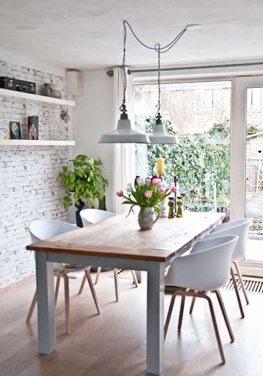 Coin repas loft avec table de cuisine en bois brut et chaises coques en plastique blancs. Suspensions loft et papier peint brique appuient cette déco de cuisine industrielle