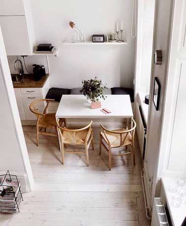Cuisine blanche avec banquette et table rectangulaire comme coin repas. Assortie avec chaises en bois vintage et coussins gris en harmonie avec les murs blancs et le plan de travail en bois.