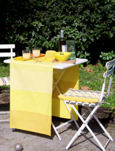 Sur la table de jardin en fer blanc, chemin de table jaune à larges rayures collection Luz Limonade. Galettes de chaise modèle Amaya. Collection Tissage de Luz