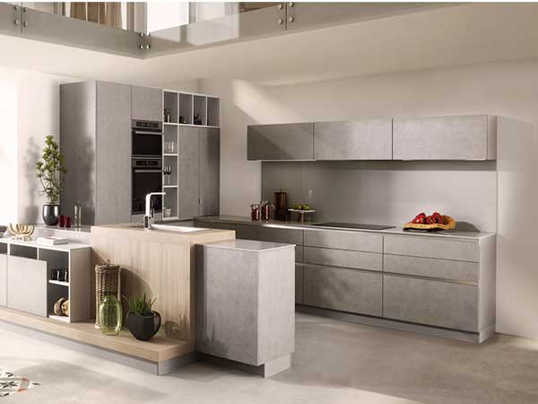 Couleurs grises minimalistes dans la cuisine américaine avec rangements intégrés, meubles de cuisines hauts et bas et évier à fleur sur plan de travail surélevé pour une ergonomie parfaite. Schmidt
