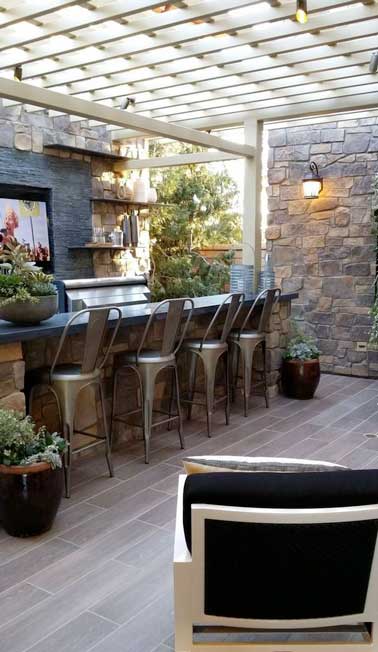 Ambiance bar chic pour la cuisine d’été aménagée sous la pergola en bois. Plan de travail en pierre naturelle, grand barbecue multifonctions et chaises de bar en alu pour la touche déco. 