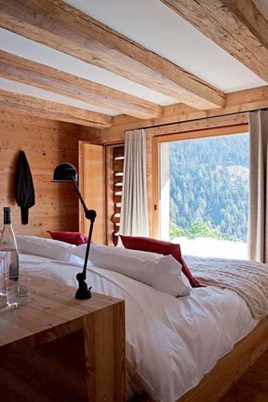 Une chambre à la déco cocooning dans un chalet de montagne. Fenêtre panoramique, lampe de chevet articulée et coussins rouges s'accordent en toute simplicité