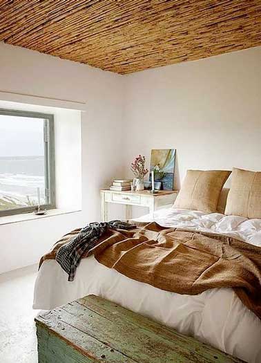 Chambre avec fenêtre panoramique vue mer et plafond tapissé en canisse. Mixé avec du linge de lit ocre et une malle en bois vieilli. Bureau vintage et murs blancs associés.