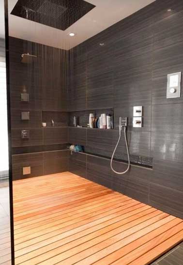 Grande douche italienne en carrelage gris avec receveur caillebotis bois. Elle combine douche pluie, colonne de douche et espace hamac. Des niches sont intégrées dans l'angle des murs