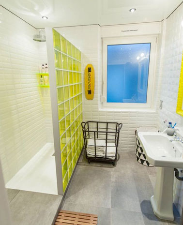 La cloison de douche italienne en carreaux de verre jaune structure la petite salle de bain éclairée par des spots encastrés dans le plafond blanc. Un mix carrelage métro dalles de béton complète l'ensemble
