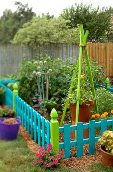 Petit jardin potager délimité par clôture en bois colorée agrémentée de pieds décoratifs. A l’intérieur des trépieds bois reçoivent une suspension pour plante grimpante.