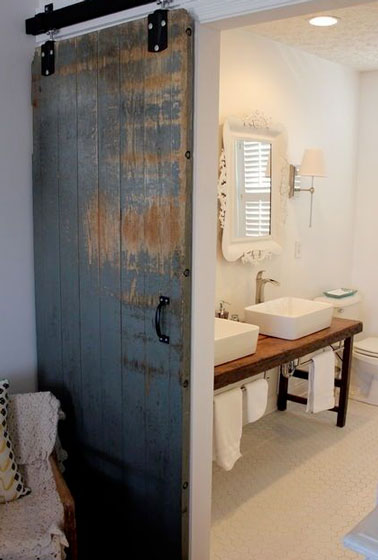 Une porte récup utilisée en cloison coulissante donne le ton de la déco authentique de cette petite salle de bain blanche. Gardée dans son jus, elle est seulement vernie