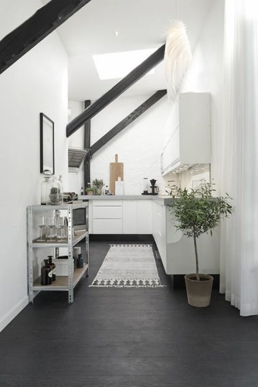 Une cuisine sous les toits, ouverte sur la pièce principale Une cuisine ouverte permet d'abattre les cloisons pour faire faire entrer plus de lumière dans un appartement sous les toits. Le blanc très lumineux contraste avec les poutres et le sol noirs.