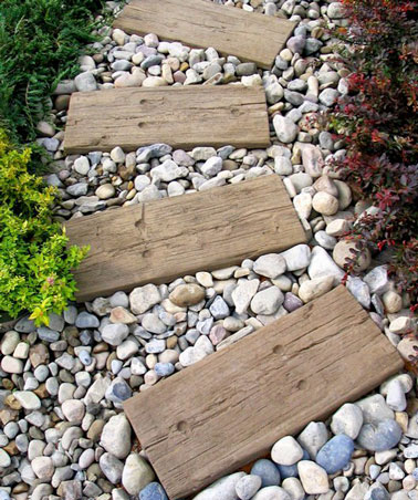 Une élégante allée de jardin fait à partir de planches de palette en bois posées sur du gravier de cailloux de divers couleurs, en accord avec les plantes autour.