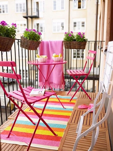 Sur ce balcon au sol en caillebotis bois clair, la couleur rose fait sa star ! Table et chaises de jardin font un clin d'oeil aux fleurs, au tapis et au plaid pour une déco vive