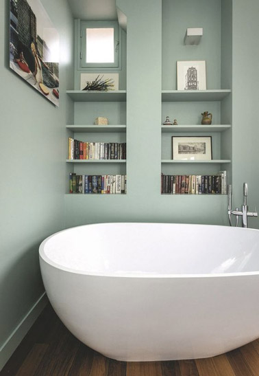 Douceur et simplicité font la pair dans cette petite salle de bain aux murs vert pastel et au sol en parquet aménagée avec une baignoire îlot et une bibliothèque ! 