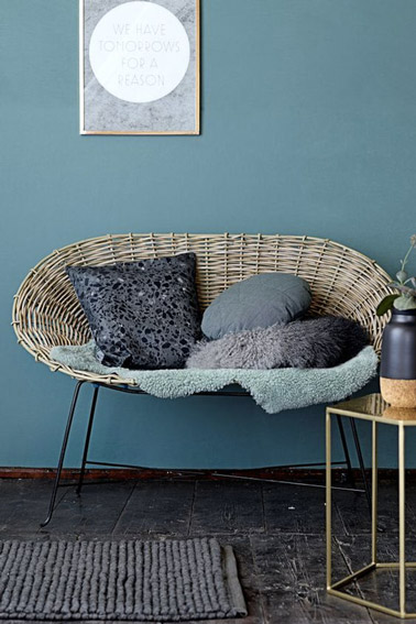 Voilà un salon de style contemporain aux murs de couleur bleu canard meublé par une table basse aux lignes géométriques et un canapé en rotin sur pieds en métal noir 