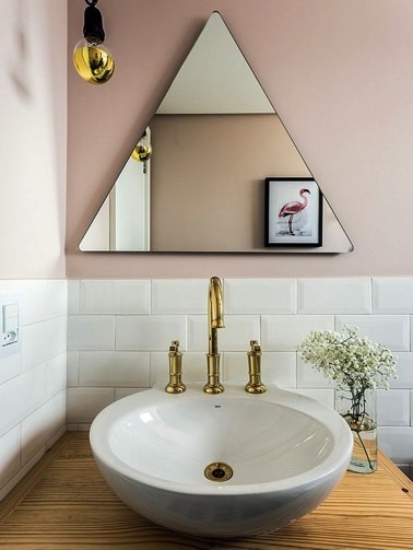 La salle de bain accueille un miroir en forme de triangle disposé sur le mur rose pastel, juste au dessus du carrelage métro blanc, le tout souligné par des touches dorées 
