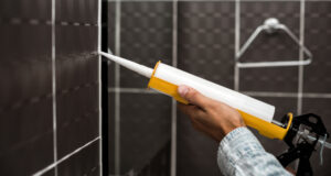 Personne posant un joint de silicone dans une salle de bain au carrelage noir avec une cartouche de silicone spécial salle de bains et d’un pistolet adapté