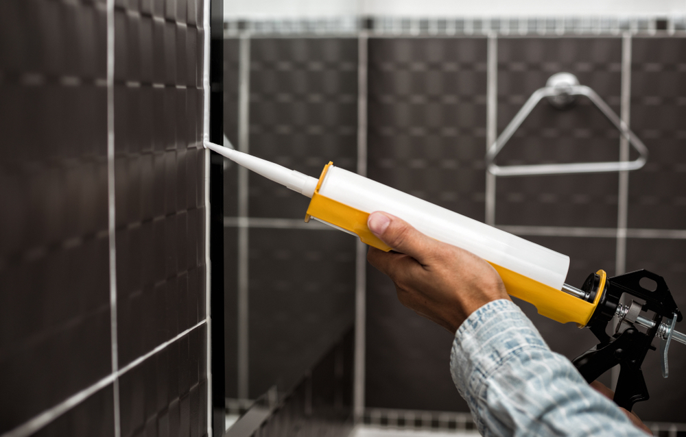 Personne posant un joint de silicone dans une salle de bain au carrelage noir avec une cartouche de silicone spécial salle de bains et d’un pistolet adapté
