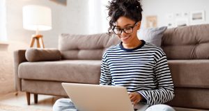 Cheerful young ethnic female freelancer in glasses and casual clothes focusing on screen and interacting with laptop while sitting alone on floor in light modern living room