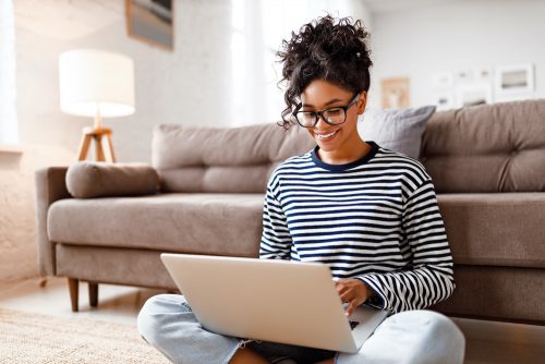 Cheerful young ethnic female freelancer in glasses and casual clothes focusing on screen and interacting with laptop while sitting alone on floor in light modern living room
