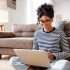 Cheerful young ethnic female freelancer in glasses and casual clothes focusing on screen and interacting with laptop while sitting alone on floor in light modern living room